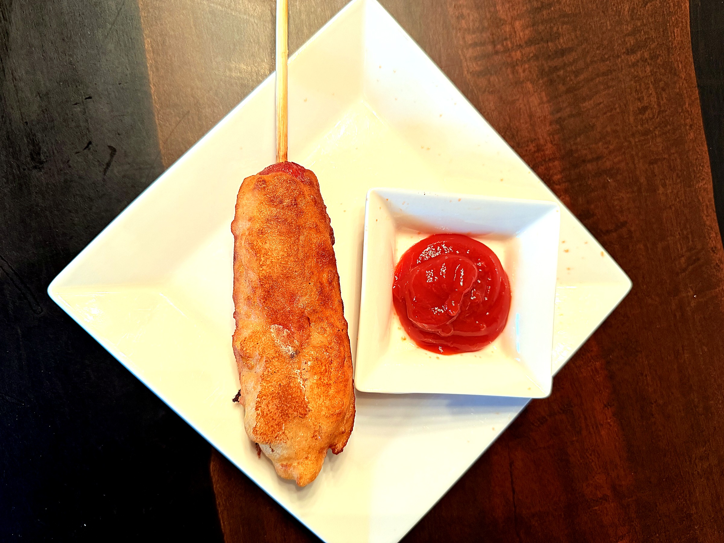 A golden fried corn dog on a stick on a white plate, next to a small dish of ketchup