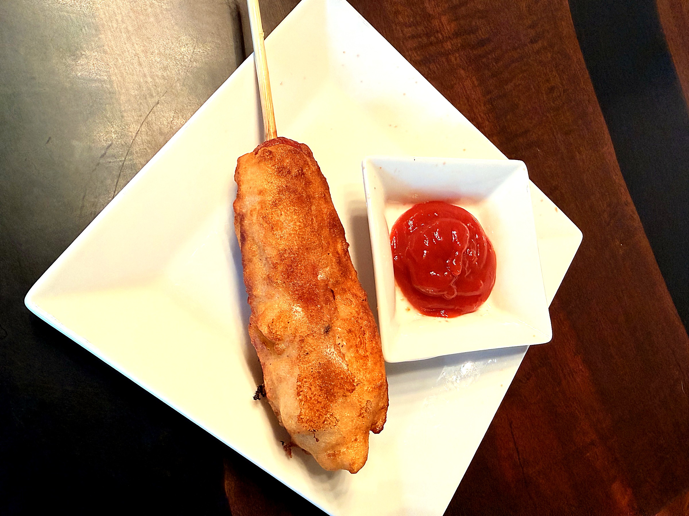 A golden fried corn dog on a stick next to a small dish of ketchup, on a white plate