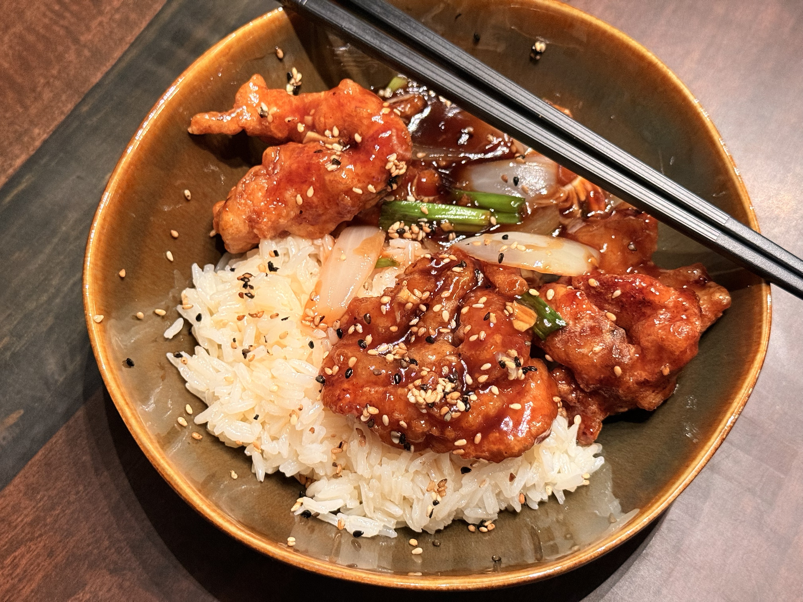 Crisp, sesame-glazed chicken over steamed rice with sautéed sweet onion and scallions in a brown bowl