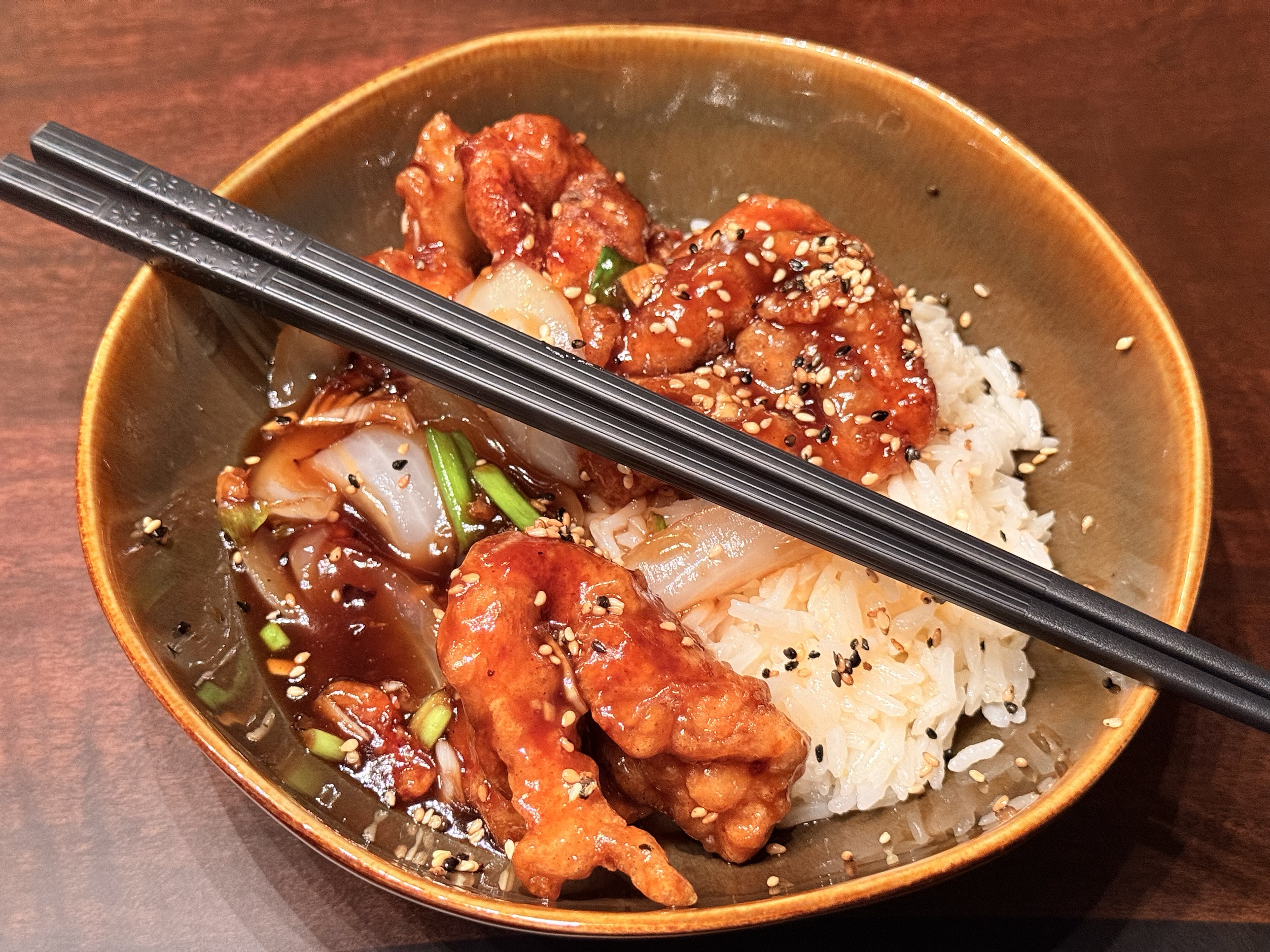Glossy, sesame-glazed chicken pieces over steamed rice in a brown bowl, with sweet onion wedges and scallions in a dark sauce