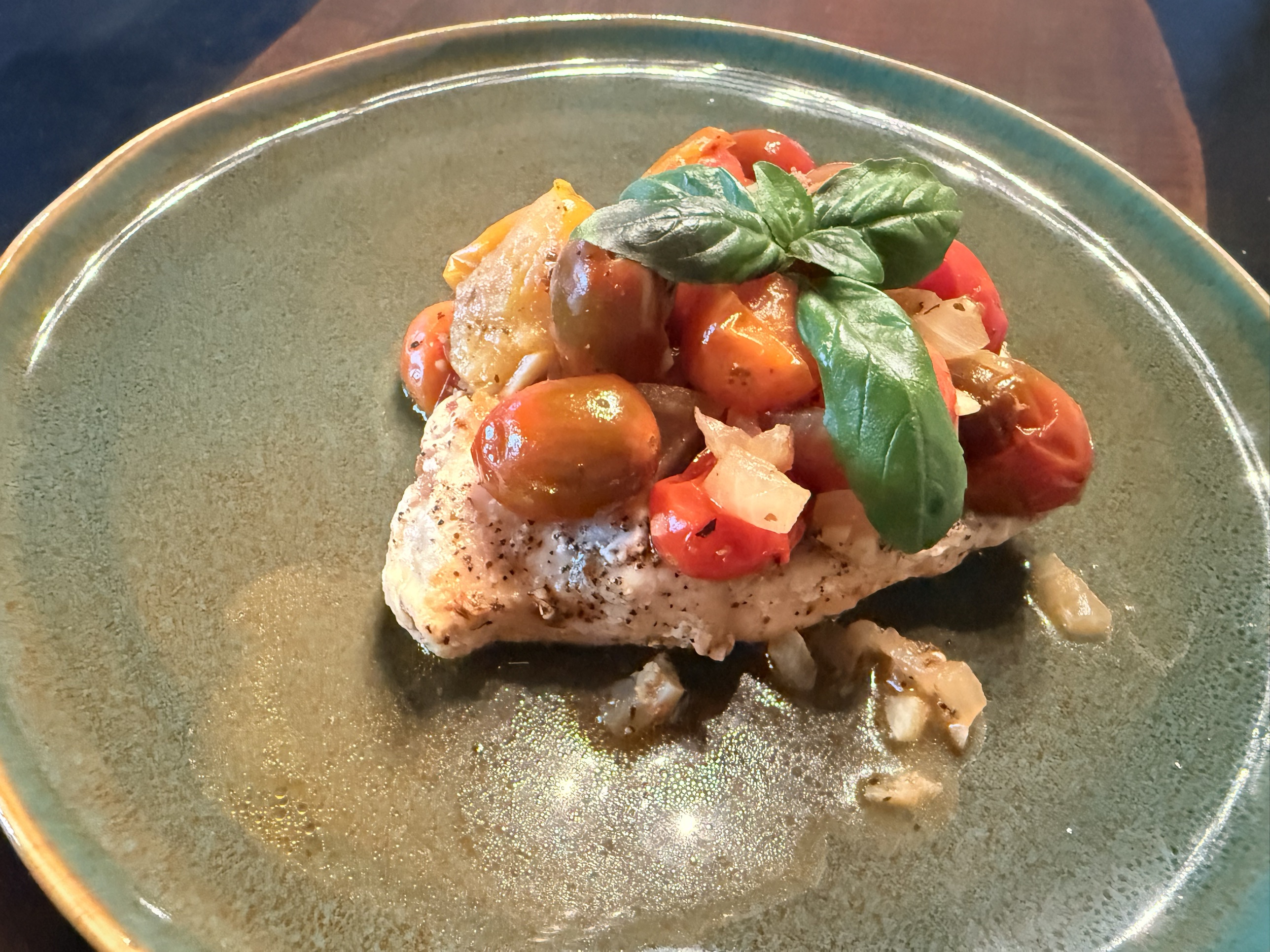 A seared chicken breast topped with burst red and golden cherry tomatoes, softened onion, and a sprig of fresh basil, sitting in a pool of lemony pan sauce on a speckled teal plate