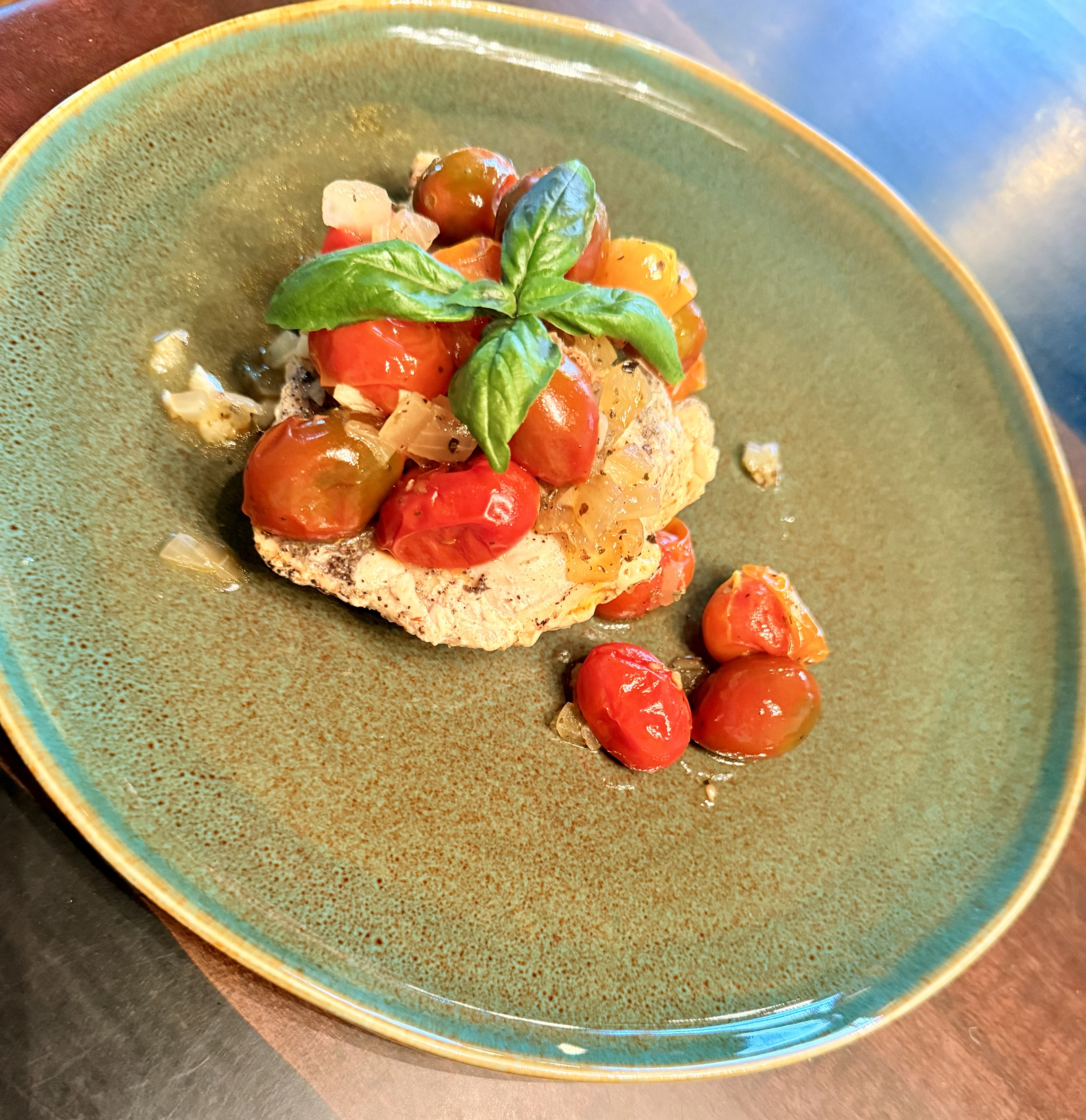 An overhead view of a seared chicken breast piled with burst cherry tomatoes and soft onions, crowned with fresh basil, on a speckled teal plate