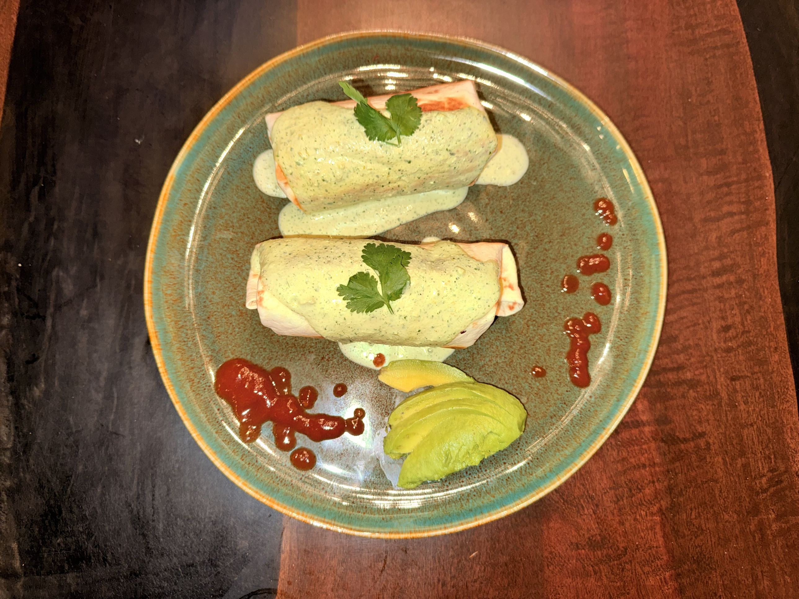 An overhead shot of two burritos smothered in a creamy green cilantro sauce and topped with cilantro leaves, served on a speckled teal plate with sliced avocado and dots of taco sauce