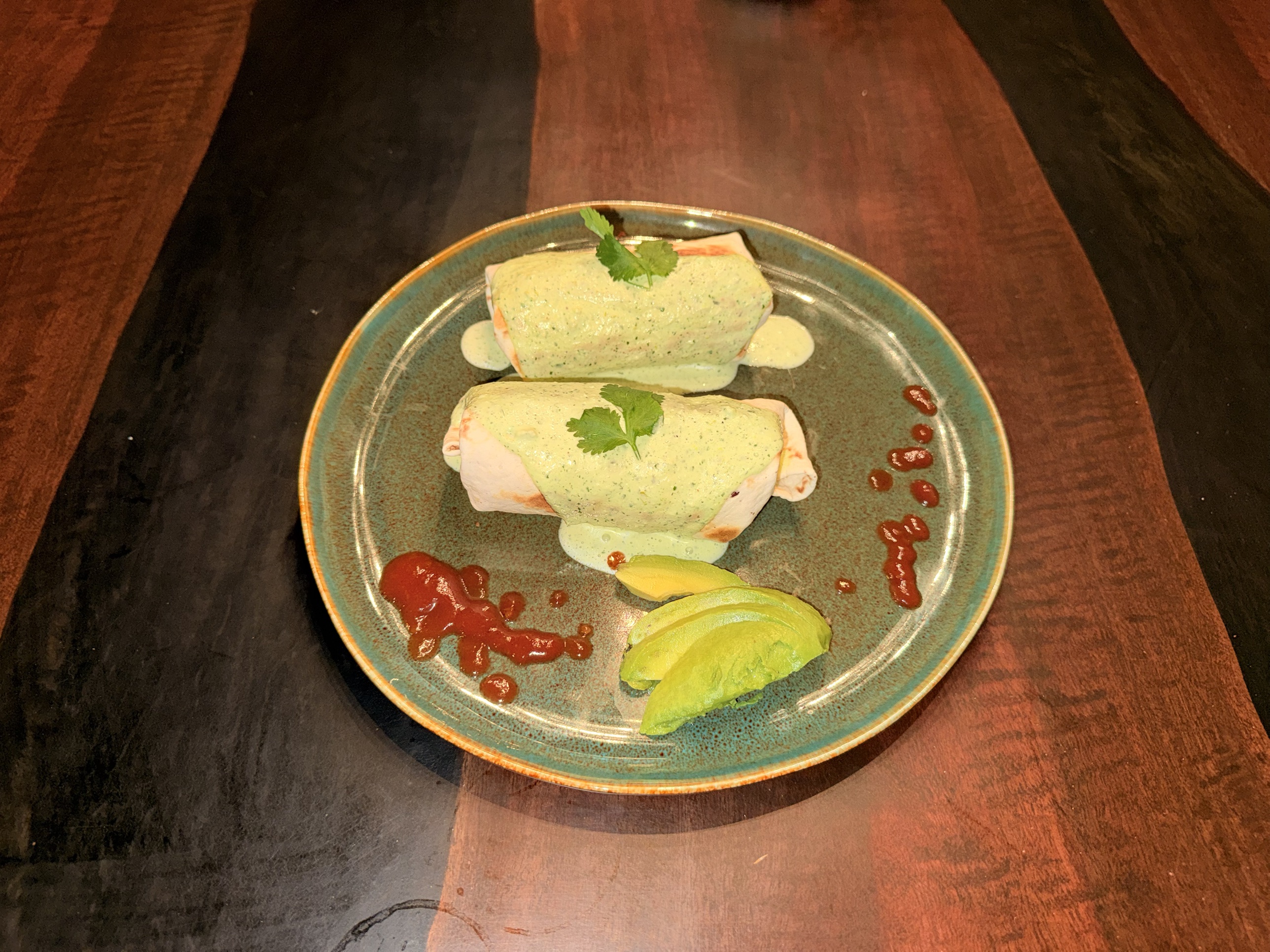 Two green-sauce smothered burritos on a speckled teal plate with avocado slices, set on a dark wood table