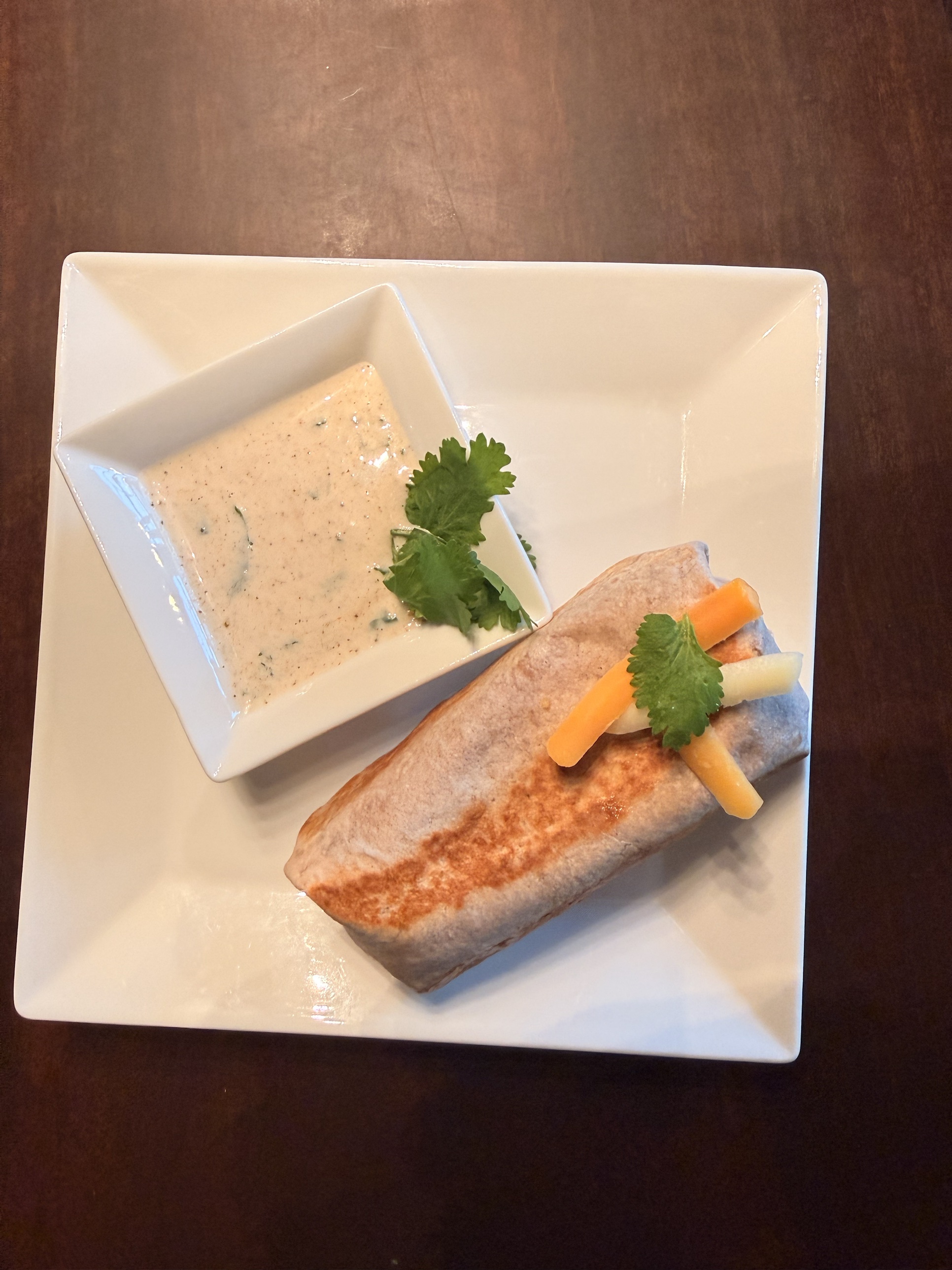 An overhead shot of a pan-crisped tortilla burrito topped with cheese and jicama sticks and cilantro, served on a square white plate next to a small dish of creamy Southwest ranch dipping sauce